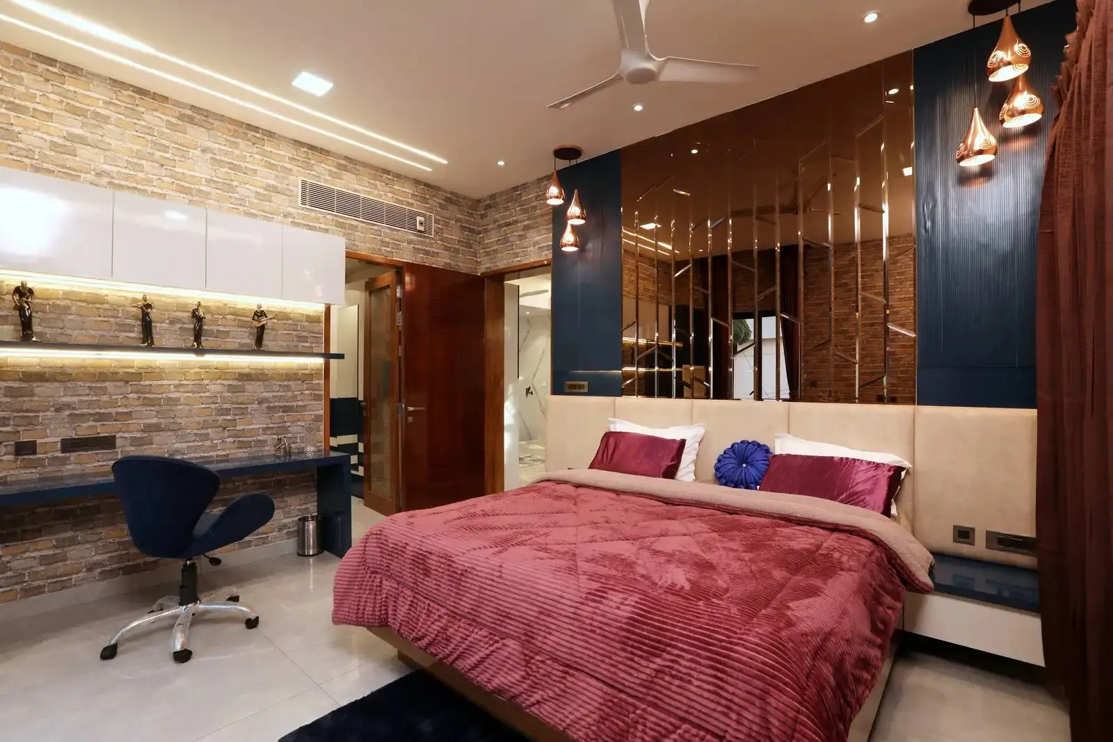 Modern bedroom with a large bed covered in a textured mauve blanket and matching pillows, a blue circular decorative pillow, brick accent wall with white cabinets and a desk with a blue swivel chair, metallic pendant lights, and a ceiling fan.