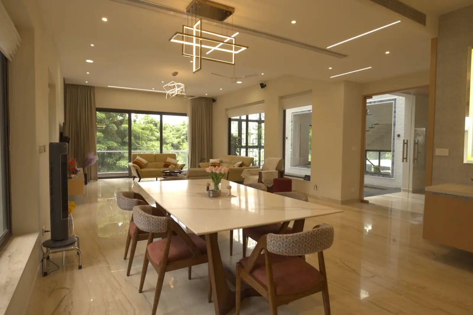 Modern dining area with a rectangular white table surrounded by six wooden chairs with patterned cushions, overlooking a living room with beige sofas and large windows with greenery outside, featuring geometric pendant lights and marble flooring.