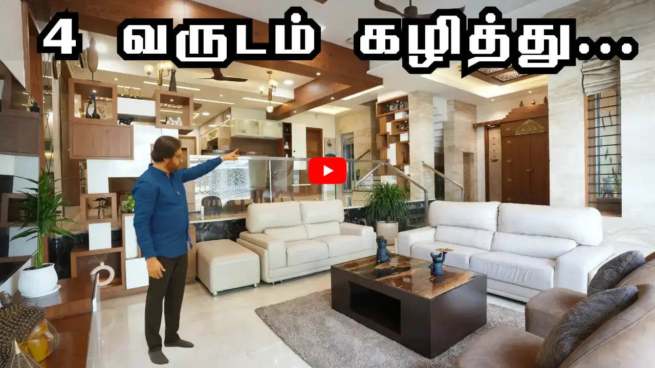 Man in a blue shirt pointing inside a spacious, modern living room with white sofas, a dark marble coffee table, and wooden shelves, with Tamil text overlay at the top.