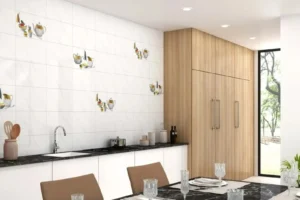 Modern kitchen with white tiled walls featuring small decorative images, white lower cabinets with a black marble countertop, wooden tall cabinets, and a dining table with brown chairs and crystal glassware.